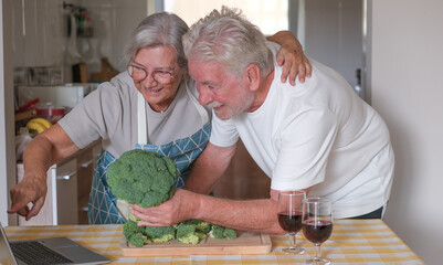 Happy older couple at the kitchen counter chopping fresh broccoli while drinking a glass of red wine and looking recipes on their laptop. Concept of preparing healthy meals and home cooking together
