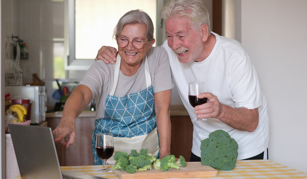 Happy older couple at the kitchen counter chopping fresh broccoli while drinking a glass of red wine and looking recipes on their laptop. Concept of preparing healthy meals and home cooking together