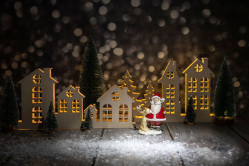 Obraz premium A miniature Christmas village, a toy Santa Claus with a reindeer on a wooden table, blurred side lights in the background, a festive holiday scene.