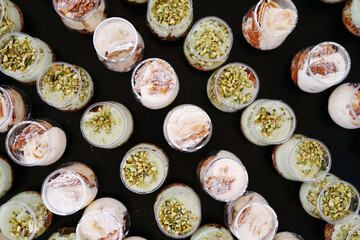 Overhead view of numerous individual dessert shots in small glass cups, featuring layered cream and pistachio garnishes, arranged randomly on a dark surface