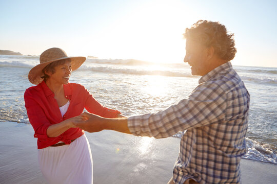 Love, mature couple and dancing on beach for romantic date, anniversary celebration and travel. Flare, happy people and holding hands with movement at seaside for summer holiday, bonding and support - Powered by Adobe