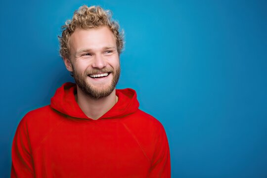 Portrait of a smiling blonde man in red hoodie against blue background, studio shot with space for text. Friendly casual male presenting approachable vibe, ideal for designs needing copy space.