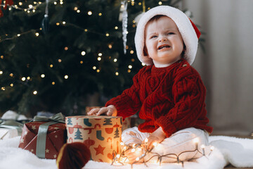 Cute baby in santa hat crying at present boxes with garland lights under modern decorated christmas tree. Sad overwhelmed baby. Holiday time
