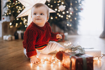 Adorable happy baby in knitted sweater sitting with stylish presents and garland lights on background of decorated christmas tree with star. Atmospheric holiday time, Merry Christmas!