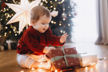 Merry Christmas and Happy Holidays! Cute baby in knitted sweater sitting with stylish gift box and...
