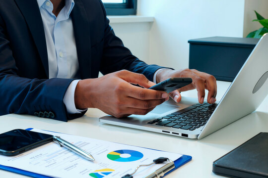 Young adult Black man working at desk using smartphone and laptop, analyzing business charts and financial documents, multitasking with technology in modern office setting