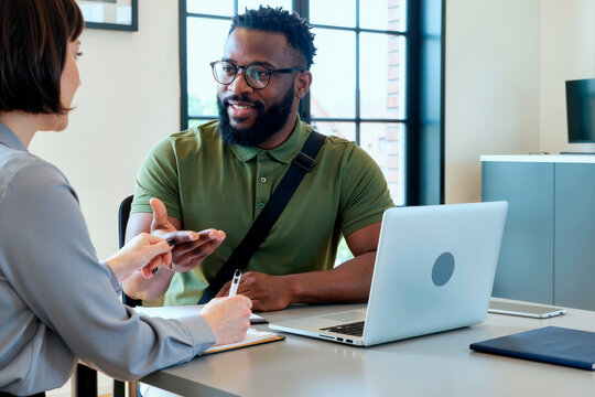 Black young adult man sitting at desk discussing business with Caucasian young adult woman, gesturing with hand while woman holding pen, open laptop and documents on table in office setting