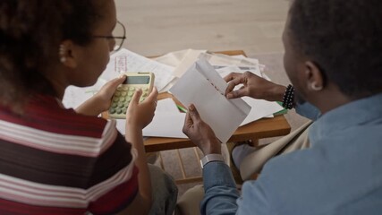 Over the shoulder shot of couple sitting at home opening envelope with overdue bill and reviewing finances together, showing concern and stress while managing household expenses - Powered by Adobe