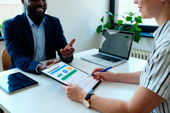 Caucasian young adult woman holding clipboard with financial chart, discussing business strategy with Black young adult man in office, both sitting at desk with laptop and tablet visible