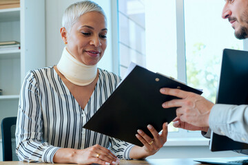 Middle aged Caucasian woman wearing neck brace sitting at desk reviewing documents with young adult Caucasian man holding clipboard during consultation in office setting