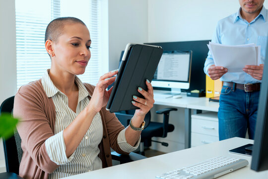 Caucasian middle aged woman using digital tablet at desk in modern office while standing Caucasian middle aged man holding documents in background, both engaged in work tasks