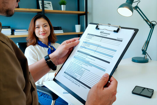 Asian young adult woman sitting at desk smiling while Caucasian young adult man holding clipboard with resume conducting job interview in modern office setting - Powered by Adobe