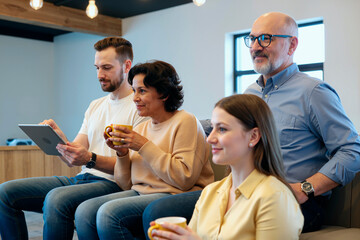 Group of middle aged and young adult Caucasian men and women sitting together, holding mugs and digital tablet participating in meeting at office workplace