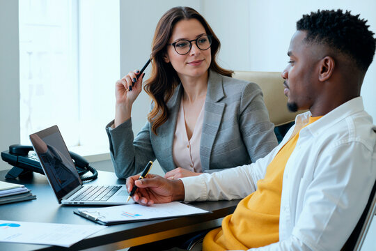 Caucasian young adult woman discussing business strategy with Black young adult man in modern office setting, both reviewing financial documents and using laptop computer