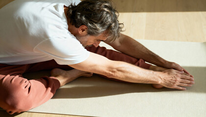 A fit middle-aged man doing a seated forward bend yoga pose on a mat. Man stretching during a morning workout in bright sunlight. Flexibility and wellness concept