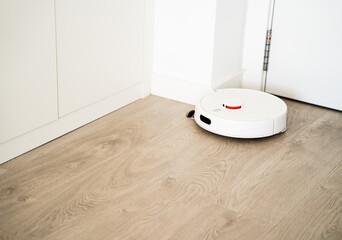 Smart robotic vacuum cleaning wooden floor near white cabinet in bright minimalist home interior environment