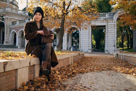 Mujer joven con abrigo de invierno hablando por tel&eacute;fono m&oacute;vil en un parque urbano de oto&ntilde;o.