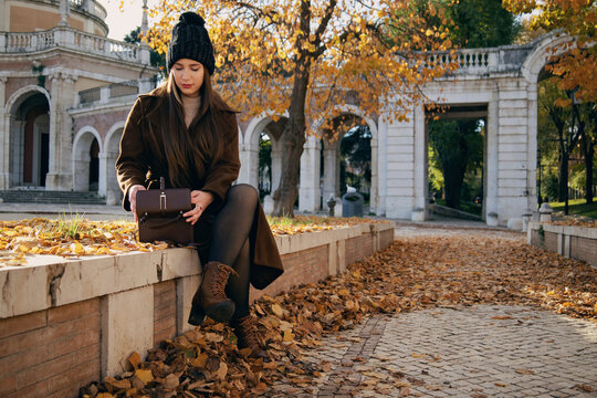 Mujer joven con abrigo marr&oacute;n y gorro de invierno en un entorno de parque con &aacute;rboles de oto&ntilde;o