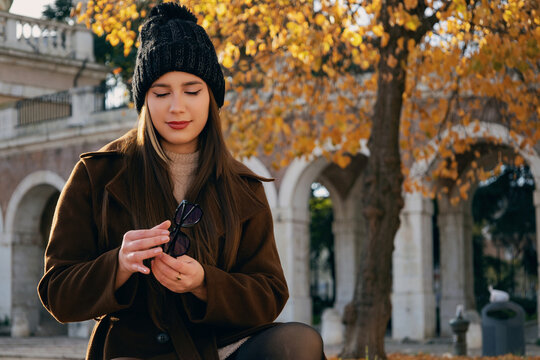 Mujer joven con abrigo marr&oacute;n y gorro de invierno en un entorno de parque con &aacute;rboles de oto&ntilde;o