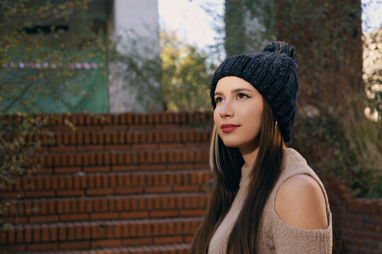 Mujer joven con gorro y bufanda de invierno posando casualmente en parque urbano soleado.