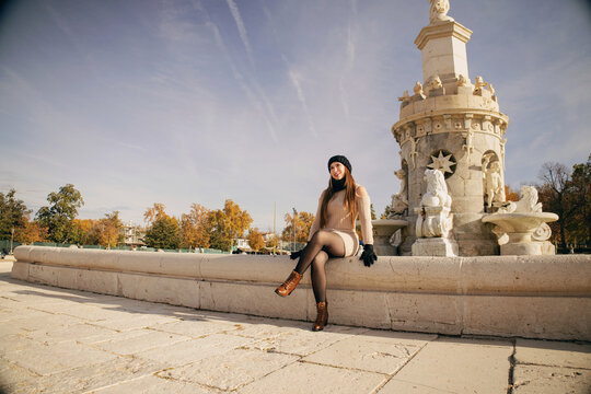 Mujer joven con gorro y bufanda de invierno posando casualmente en parque urbano soleado.