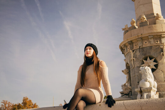 Mujer joven con gorro y bufanda de invierno posando casualmente en parque urbano soleado.