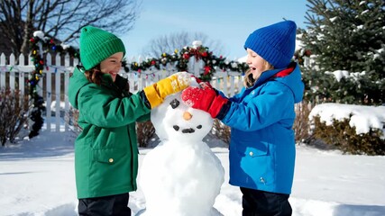 Two children in winter coats build a snowman together on a sunny, snowy day