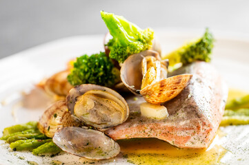 Close-up of gourmet pan-seared salmon fillet topped with clams, broccoli, and asparagus in a white wine or butter sauce on a white plate, served in a restaurant setting