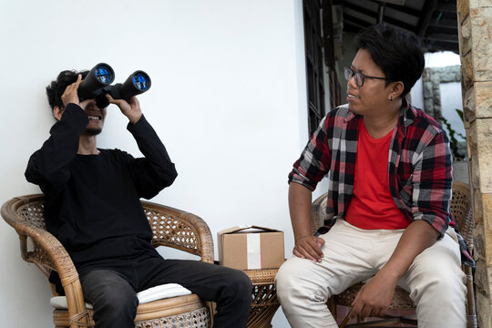 Two Indonesian southeast asian men sitting on a chair. Showing a package of a new pair of binoculars to his friend. Delivery service