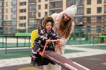 A mother helps her young child play on a seesaw at a city park. The child wears a warm coat and struggles with a frown while having fun