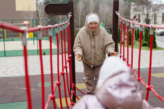 A senior woman engages playfully with a young child on a bridge-like structure at a colorful playground during a cold afternoon