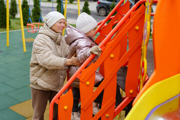 An elderly woman helps a young girl ascend the steps of a vibrant playground structure during a clear, sunny day, showcasing a moment of care and joy