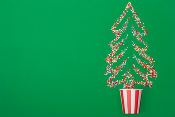 Multicolored sugar stars in the shape of a Christmas tree are scattered across a green background. A paper cup is also placed on the green background. Viewed from above, flat lay