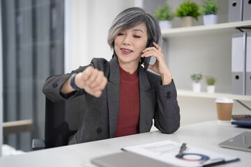 Asian older adult businesswoman check smartwatch talking mobile phone modern office busy professional setting time management communication work