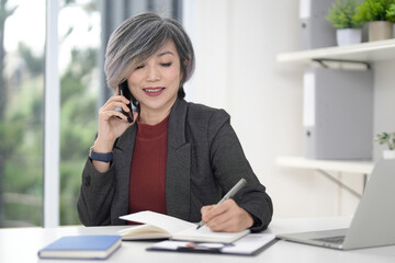 Asian older businesswoman professional phone call writing notes office desk consults clients manages business operations
