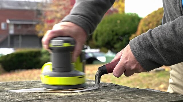 Focus on a person sanding a masonry trowel outside as fall leaves provide a colorful backdrop. The person carefully smooths the metal, preparing it for future use.