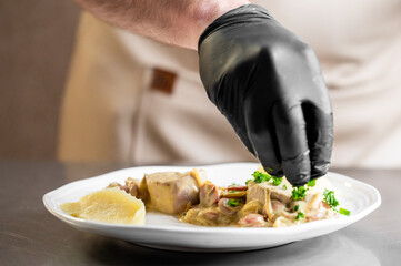 A chef wearing black gloves is adding the final garnish of fresh parsley to a gourmet dish of meat and mushrooms in a creamy sauce, served with a side of mashed potatoes on a white plate 