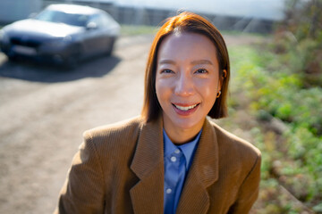 Woman smiling joyfully outdoors in a sunny setting near a parked car surrounded by greenery