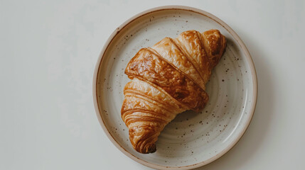 A golden brown, flaky croissant resting on a rustic, speckled ceramic plate against a plain, light background