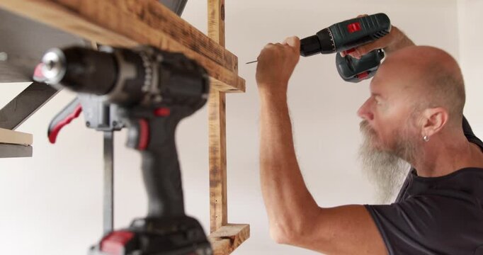 50 year craftsman assembling wooden shelf at home, fixing slates using wood screws and electric screwdriver, real people
