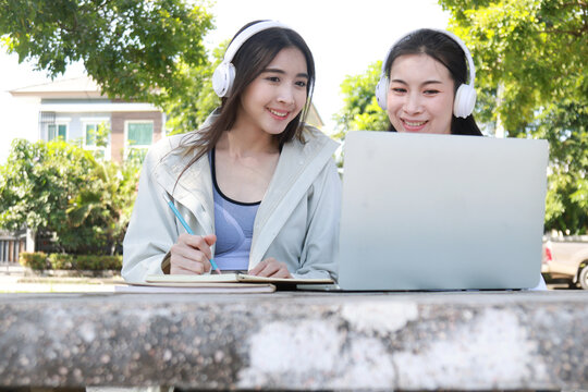Pretty cheerful asian woman in casual clothes browses laptop computer connected to 4g internet updates software uses modern technologies poses outdoor looks gladfully away with her friend. 