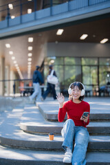 Fototapeta premium Asian student wearing headphones waving sitting on campus steps