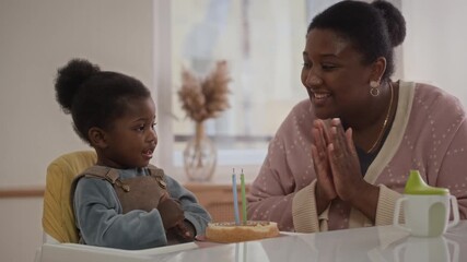 Loving Black mother celebrating birthday of her two-year-old daughter, blowing candles on cake and clapping hands