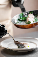 A chef wearing black gloves carefully uses a metal spoon to plate a gourmet dish with sauce on a white plate; shallow depth of field, natural lighting