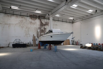 The interior of a large, with high ceilings and white metal beams. In the center, a medium-sized motorboat rests on metal trestles that lift it off the ground for maintenance or storage.