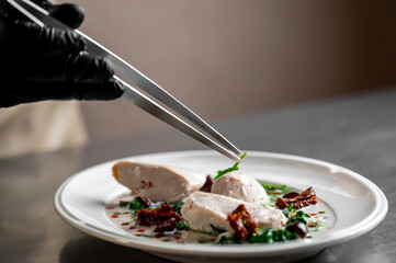 A chef in black gloves uses tweezers to meticulously place a green garnish on a gourmet chicken dish with sun-dried tomatoes and spinach in a creamy sauce.
