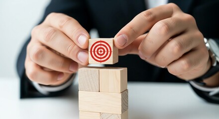A close-up shot shows a businessman's hands carefully placing a wooden block with a red and white bullseye target symbol onto a stack of plain wooden blocks. The focus is sharp on the target block,