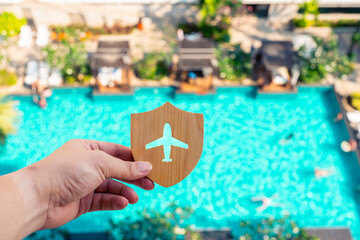 Hand holding wooden shield with airplane cutout over luxury resort pool background. Concept of travel insurance, vacation safety, secure holidays, and worry-free tourism experience.