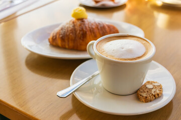 Taza de café con leche caliente y galleta tipo bizcocho sobre plato blanco, con medialuna borrosa al fondo en una mesa de cafetería iluminada.