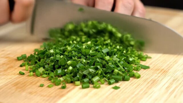 A close-up view of a hand chopping green chives on a wooden cutting board with a knife